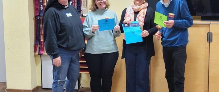Vier Personen stehen nebeneinander im Bürgerzentrum Deutz und lächeln in die Kamera. Von links nach rechts: Sarah M. als Toastmasterin des Abends, Sarah B. mit der Urkunde für die beste Bewertungsrede, Ana G. mit der Urkunde für die beste vorbereitete Rede und ihre allererste Rede überhaupt, sowie Christian B., der als Chief Judge der Mitgliederversammlung den „Hammer des Abends“ hält. Alle tragen Namensschilder, hinter ihnen hängen farbige Toastmasters-Banner. Die Kölner Sprechbar stellt mit einem Augenzwinkern fest: Es gibt eindeutig viel zu wenige Sarahs im Club.