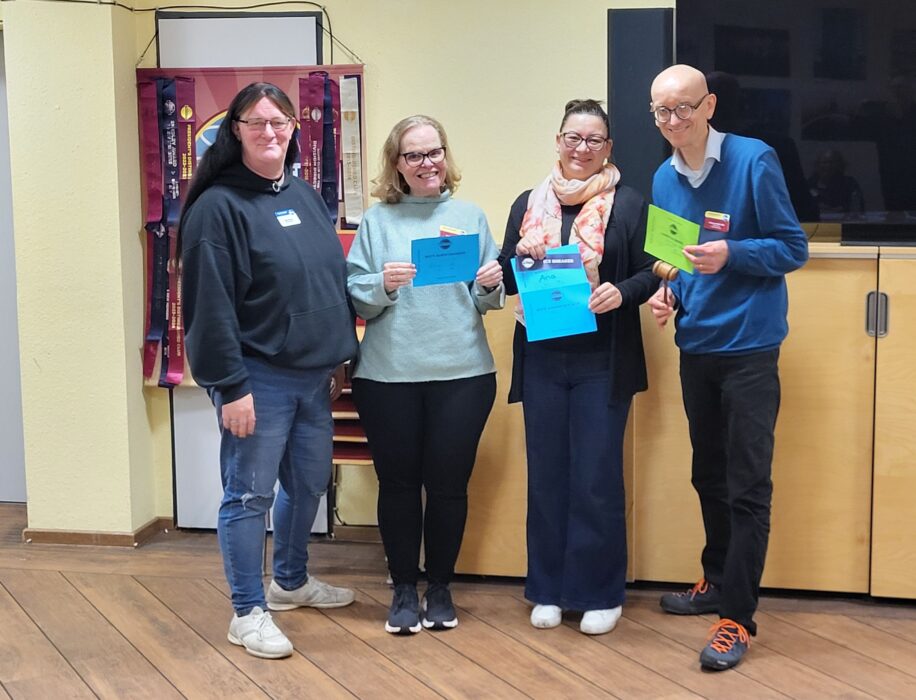 Vier Personen stehen nebeneinander im Bürgerzentrum Deutz und lächeln in die Kamera. Von links nach rechts: Sarah M. als Toastmasterin des Abends, Sarah B. mit der Urkunde für die beste Bewertungsrede, Ana G. mit der Urkunde für die beste vorbereitete Rede und ihre allererste Rede überhaupt, sowie Christian B., der als Chief Judge der Mitgliederversammlung den „Hammer des Abends“ hält. Alle tragen Namensschilder, hinter ihnen hängen farbige Toastmasters-Banner. Die Kölner Sprechbar stellt mit einem Augenzwinkern fest: Es gibt eindeutig viel zu wenige Sarahs im Club.
