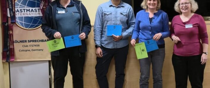 Vier Personen stehen nebeneinander in einem Raum und lächeln in die Kamera. Drei halten farbige Umschläge in Blau und Grün. Links steht ein Toastmasters-Banner mit Logo und der Aufschrift „Kölner Sprechbar“. Im Hintergrund ist ein großer schwarzer Bildschirm. Holzboden und helle Wand.