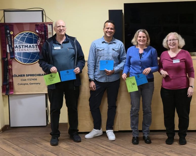 Vier Personen stehen nebeneinander in einem Raum und lächeln in die Kamera. Drei halten farbige Umschläge in Blau und Grün. Links steht ein Toastmasters-Banner mit Logo und der Aufschrift „Kölner Sprechbar“. Im Hintergrund ist ein großer schwarzer Bildschirm. Holzboden und helle Wand.