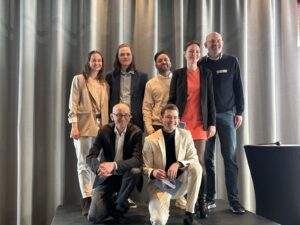 Sieben Personen posieren auf einer kleinen Bühne vor grauen Vorhängen. Zwei sitzen vorne, fünf stehen dahinter. Einige lächeln in die Kamera, eine Person hält eine Toastmasters-Mappe in der Hand.