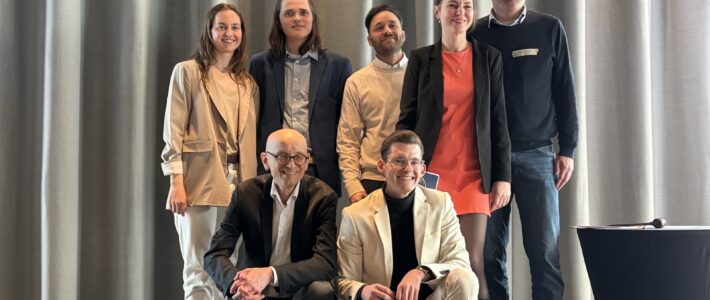 Sieben Personen posieren auf einer kleinen Bühne vor grauen Vorhängen. Zwei sitzen vorne, fünf stehen dahinter. Einige lächeln in die Kamera, eine Person hält eine Toastmasters-Mappe in der Hand.