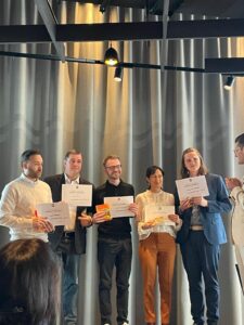 Sechs Personen stehen auf einer Bühne vor grauen Vorhängen. Fünf halten Urkunden, einige zusätzlich kleine Preise. Rechts spricht eine weitere Person zu der Gruppe. Im Vordergrund sind Köpfe von Zuschauenden zu sehen, oben schwarze Deckenstreben und Lampen.