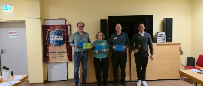 Vier Personen stehen in einem Raum vor einem Bildschirm und halten farbige Urkunden. Neben ihnen ist ein Toastmasters-Banner zu sehen. Eine Person rechts hält einen kleinen Hammer, der Raum hat helle Wände und Holzboden.