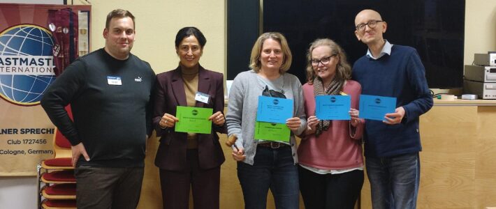 Fünf Personen stehen in einem Veranstaltungsraum nebeneinander, lächeln in die Kamera und halten farbige Urkunden. Links ist ein Toastmasters-Banner zu sehen, hinter ihnen ein großer Bildschirm und Holzmöbel. Eine Person hält zusätzlich einen kleinen Hammer in der Hand.