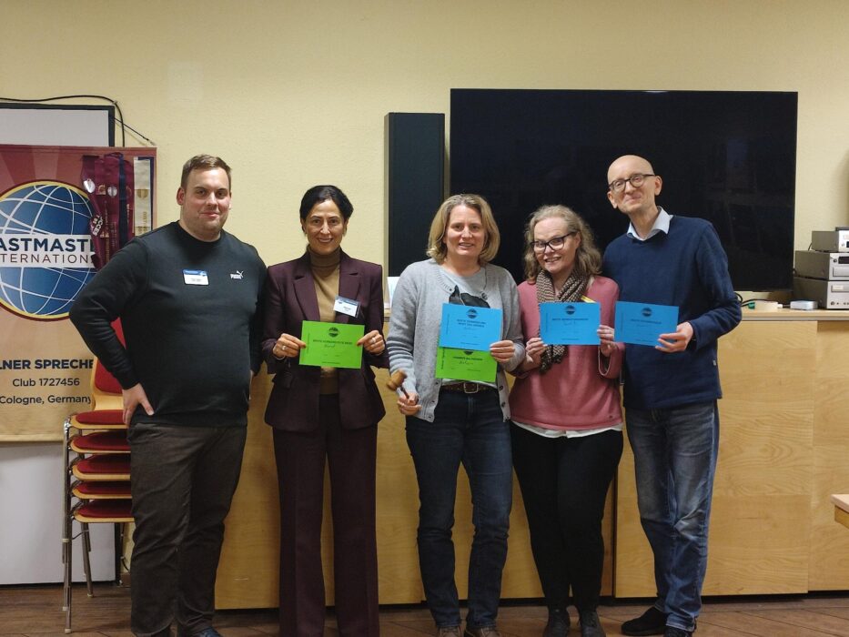 Fünf Personen stehen in einem Veranstaltungsraum nebeneinander, lächeln in die Kamera und halten farbige Urkunden. Links ist ein Toastmasters-Banner zu sehen, hinter ihnen ein großer Bildschirm und Holzmöbel. Eine Person hält zusätzlich einen kleinen Hammer in der Hand.
