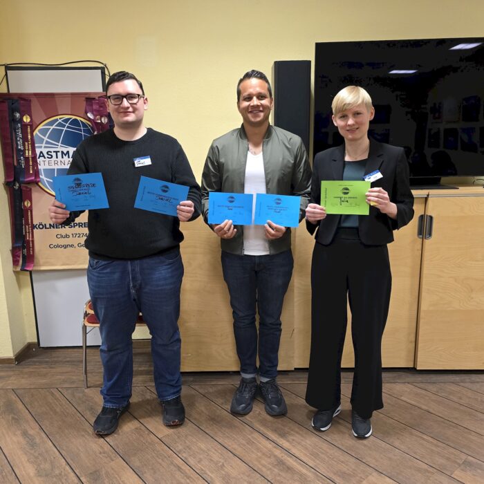 Drei Personen stehen nebeneinander in einem Raum und halten Urkunden in den Händen. Hinter ihnen befinden sich ein großer Bildschirm und ein Toastmasters-Banner. Der Raum hat helle Wände, Holzboden und wirkt wie ein Veranstaltungsraum.