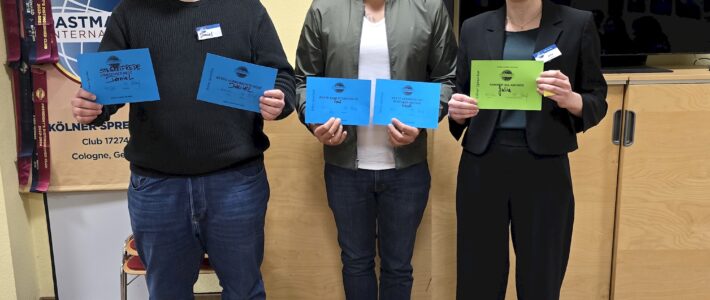 Kölner Sprechbar | Rückblick: Clubabend vom 08.04.2026 Drei Personen stehen nebeneinander in einem Raum und halten Urkunden in den Händen. Hinter ihnen befinden sich ein großer Bildschirm und ein Toastmasters-Banner. Der Raum hat helle Wände, Holzboden und wirkt wie ein Veranstaltungsraum.