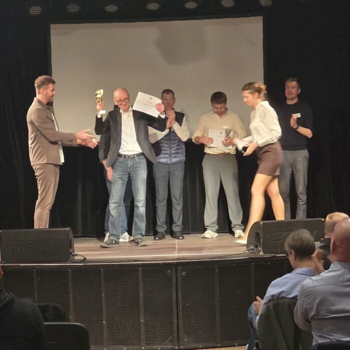 Auf einer Bühne erhält eine Person Pokal und Urkunde. Weitere Teilnehmende stehen daneben, einige applaudieren. Im Vordergrund ist Publikum zu sehen. Die Szene wirkt wie eine Preisverleihung bei einem Redewettbewerb.