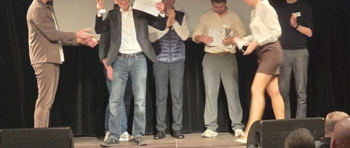 Auf einer Bühne erhält eine Person Pokal und Urkunde. Weitere Teilnehmende stehen daneben, einige applaudieren. Im Vordergrund ist Publikum zu sehen. Die Szene wirkt wie eine Preisverleihung bei einem Redewettbewerb.