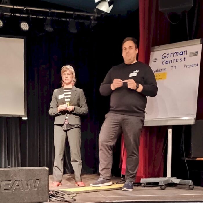 Zwei Personen stehen auf einer Bühne vor einem Flipchart mit der Aufschrift „German Contest“. Eine Person spricht vor Publikum, die andere hält Unterlagen. Bühnenlicht und Vorhang prägen den Hintergrund.