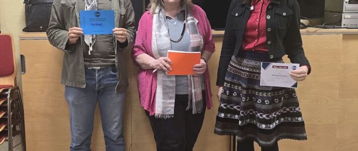 Drei erwachsene Personen stehen lächelnd vor einem großen schwarzen Bildschirm in einem Seminarraum. Zwei halten Auszeichnungen in den Händen, eine Person hält Moderationskarten. Im Hintergrund stehen Schränke, Technikgeräte und gestapelte Stühle.