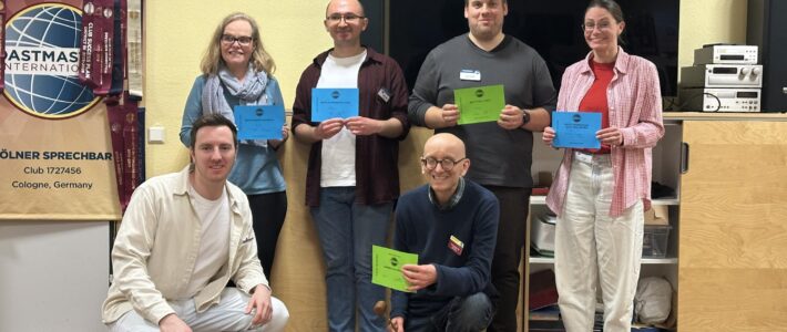 Sechs Personen posieren in einem Veranstaltungsraum vor einem großen Bildschirm und einem Toastmasters-Banner. Vier stehen, zwei knien vorne. Mehrere halten farbige Urkunden, eine Person hält zusätzlich einen kleinen Hammer. Der Raum hat helle Wände und Holzboden.