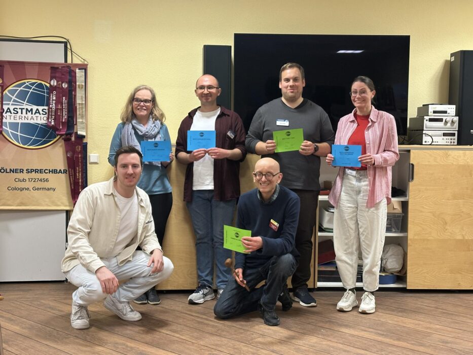 Sechs Personen posieren in einem Veranstaltungsraum vor einem großen Bildschirm und einem Toastmasters-Banner. Vier stehen, zwei knien vorne. Mehrere halten farbige Urkunden, eine Person hält zusätzlich einen kleinen Hammer. Der Raum hat helle Wände und Holzboden.