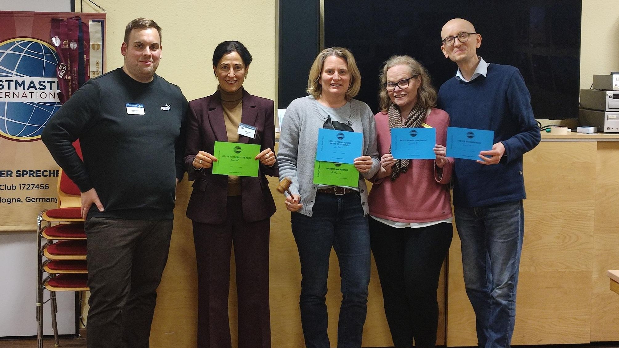 Fünf Personen stehen in einem Veranstaltungsraum nebeneinander, lächeln in die Kamera und halten farbige Urkunden. Links ist ein Toastmasters-Banner zu sehen, hinter ihnen ein großer Bildschirm und Holzmöbel. Eine Person hält zusätzlich einen kleinen Hammer in der Hand.
