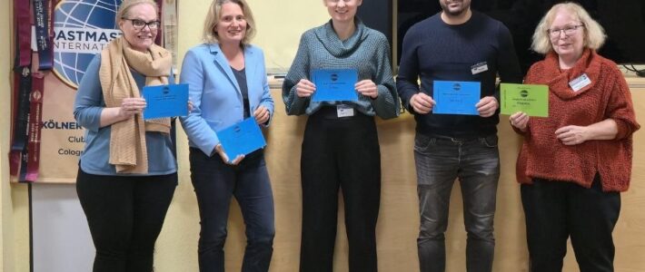 Fünf Mitglieder der Kölner Sprechbar stehen mit ihren Urkunden vor dem Toastmasters-Banner und lächeln in die Kamera.