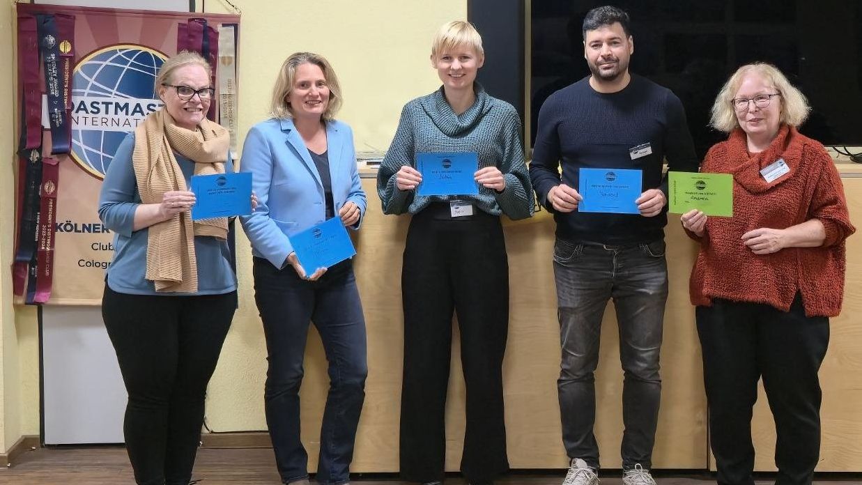 Fünf Mitglieder der Kölner Sprechbar stehen mit ihren Urkunden vor dem Toastmasters-Banner und lächeln in die Kamera.