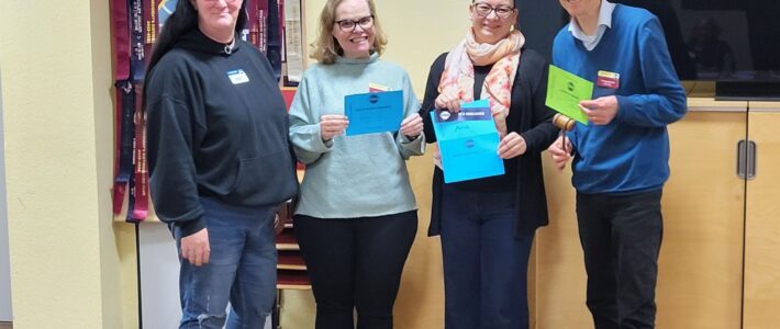 Vier Mitglieder der Kölner Sprechbar stehen mit ihren Urkunden vor dem Toastmasters-Banner und lächeln in die Kamera.