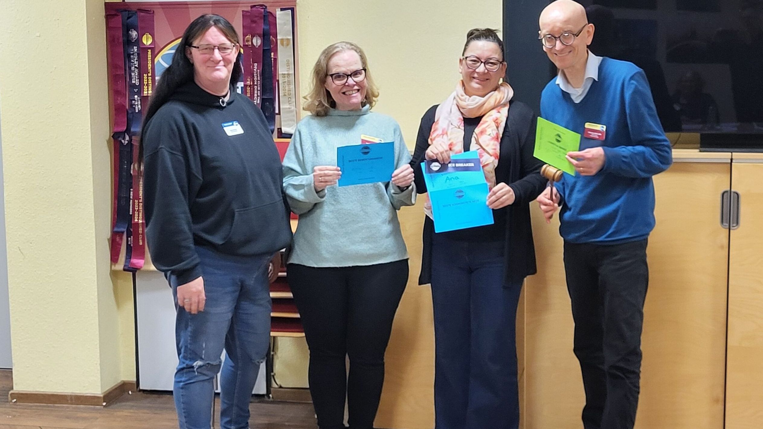 Vier Mitglieder der Kölner Sprechbar stehen mit ihren Urkunden vor dem Toastmasters-Banner und lächeln in die Kamera.