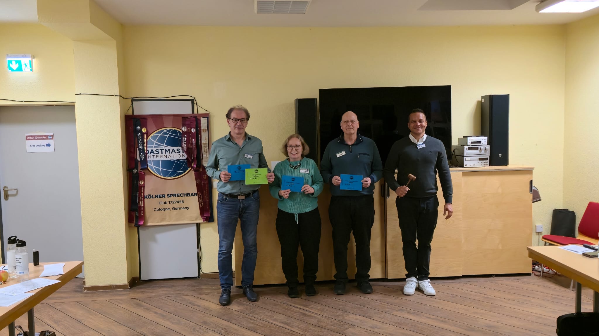 Vier Personen stehen in einem Raum vor einem Bildschirm und halten farbige Urkunden. Neben ihnen ist ein Toastmasters-Banner zu sehen. Eine Person rechts hält einen kleinen Hammer, der Raum hat helle Wände und Holzboden.