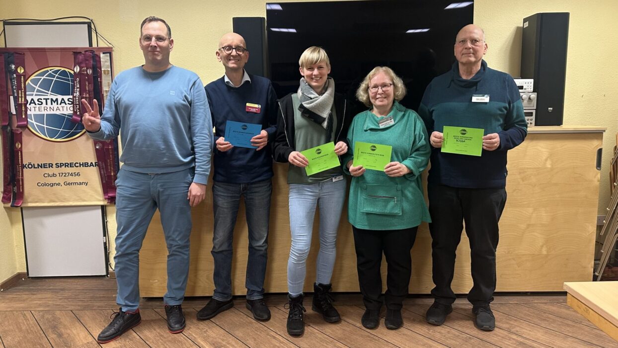 Fünf Mitglieder der Kölner Sprechbar stehen mit ihren Urkunden vor dem Toastmasters-Banner und lächeln in die Kamera.