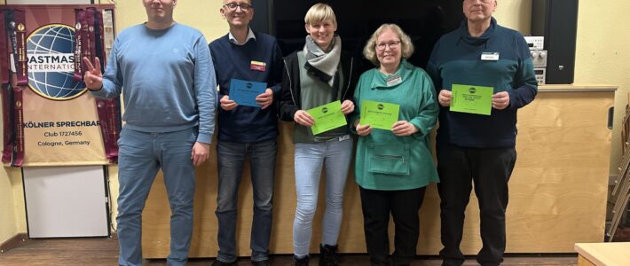 Fünf Mitglieder der Kölner Sprechbar stehen mit ihren Urkunden vor dem Toastmasters-Banner und lächeln in die Kamera.