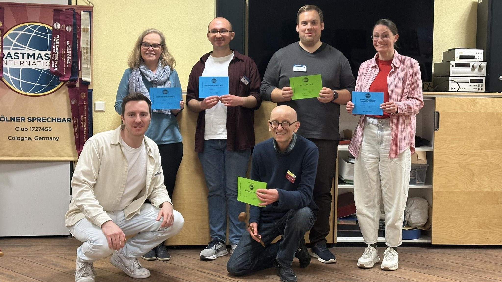 Sechs Personen posieren in einem Veranstaltungsraum vor einem großen Bildschirm und einem Toastmasters-Banner. Vier stehen, zwei knien vorne. Mehrere halten farbige Urkunden, eine Person hält zusätzlich einen kleinen Hammer. Der Raum hat helle Wände und Holzboden.