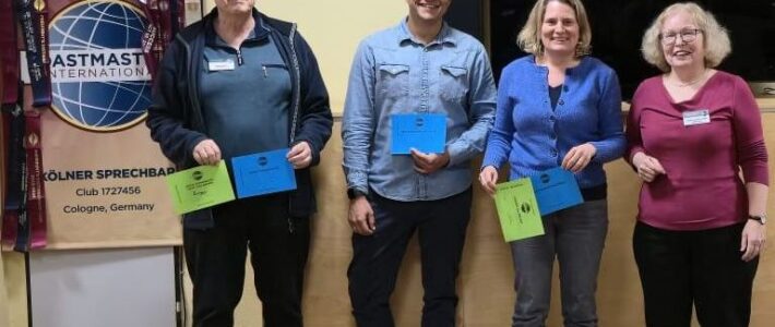 Vier Mitglieder der Kölner Sprechbar stehen mit ihren Urkunden vor dem Toastmasters-Banner; im Hintergrund ein Bildschirm und eine Holzwand.