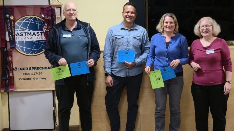 Vier Personen stehen nebeneinander in einem Raum und lächeln in die Kamera. Drei halten farbige Umschläge in Blau und Grün. Links steht ein Toastmasters-Banner mit Logo und der Aufschrift „Kölner Sprechbar“. Im Hintergrund ist ein großer schwarzer Bildschirm. Holzboden und helle Wand.