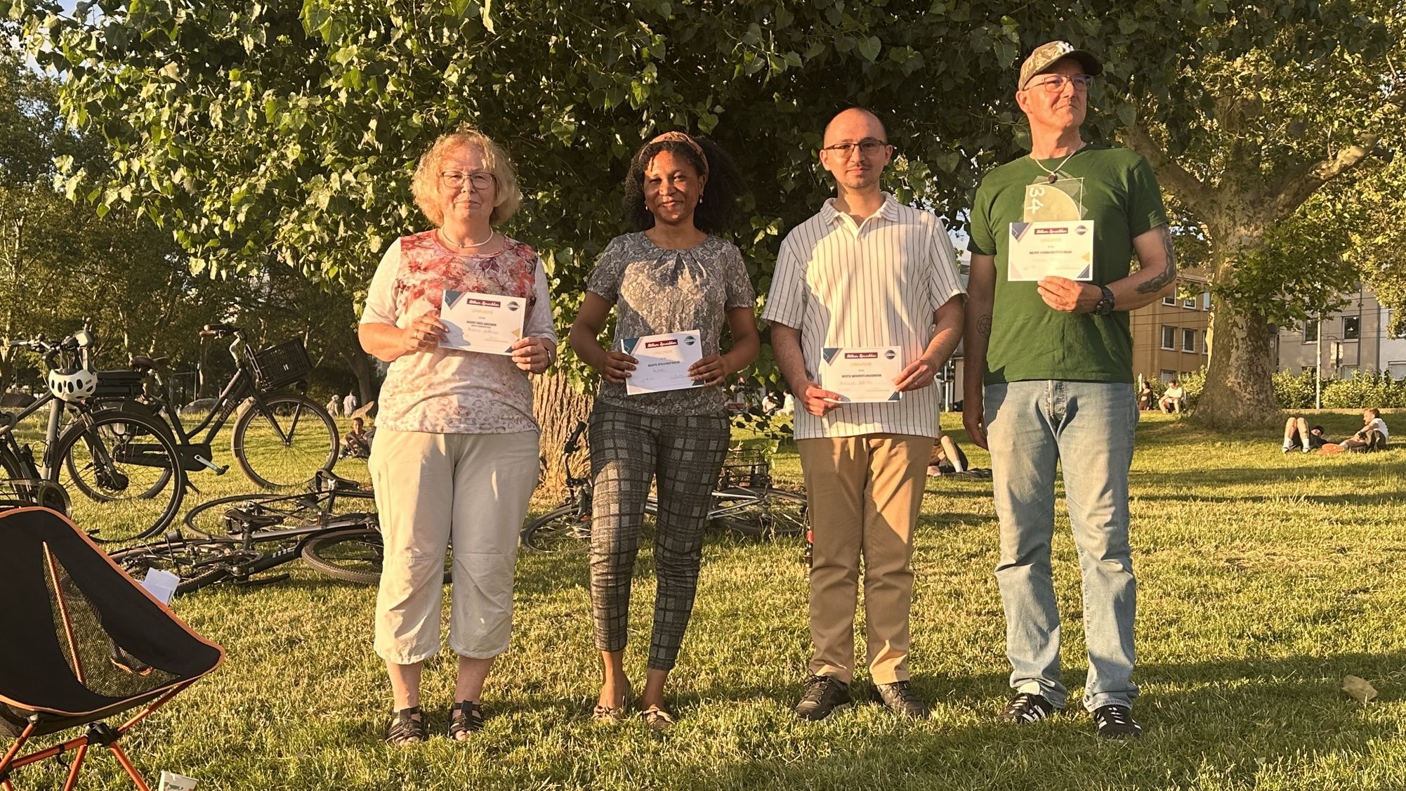Vier Mitglieder der Kölner Sprechbar präsentieren beim Draußen-Meeting am Rheinufer ihre Urkunden.