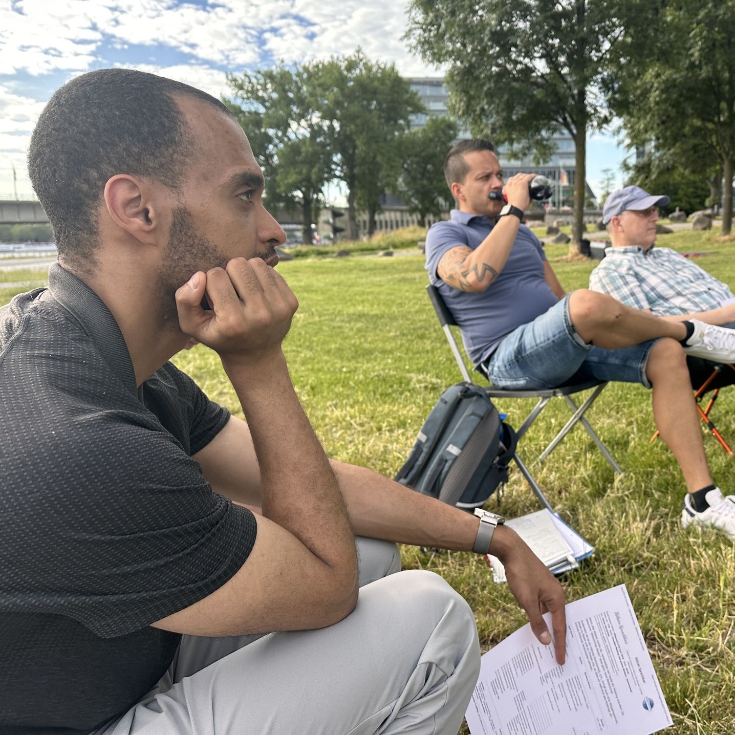 Drei Teilnehmer verfolgen auf einer Wiese am Rhein aufmerksam das Draußen-Meeting der Kölner Sprechbar.