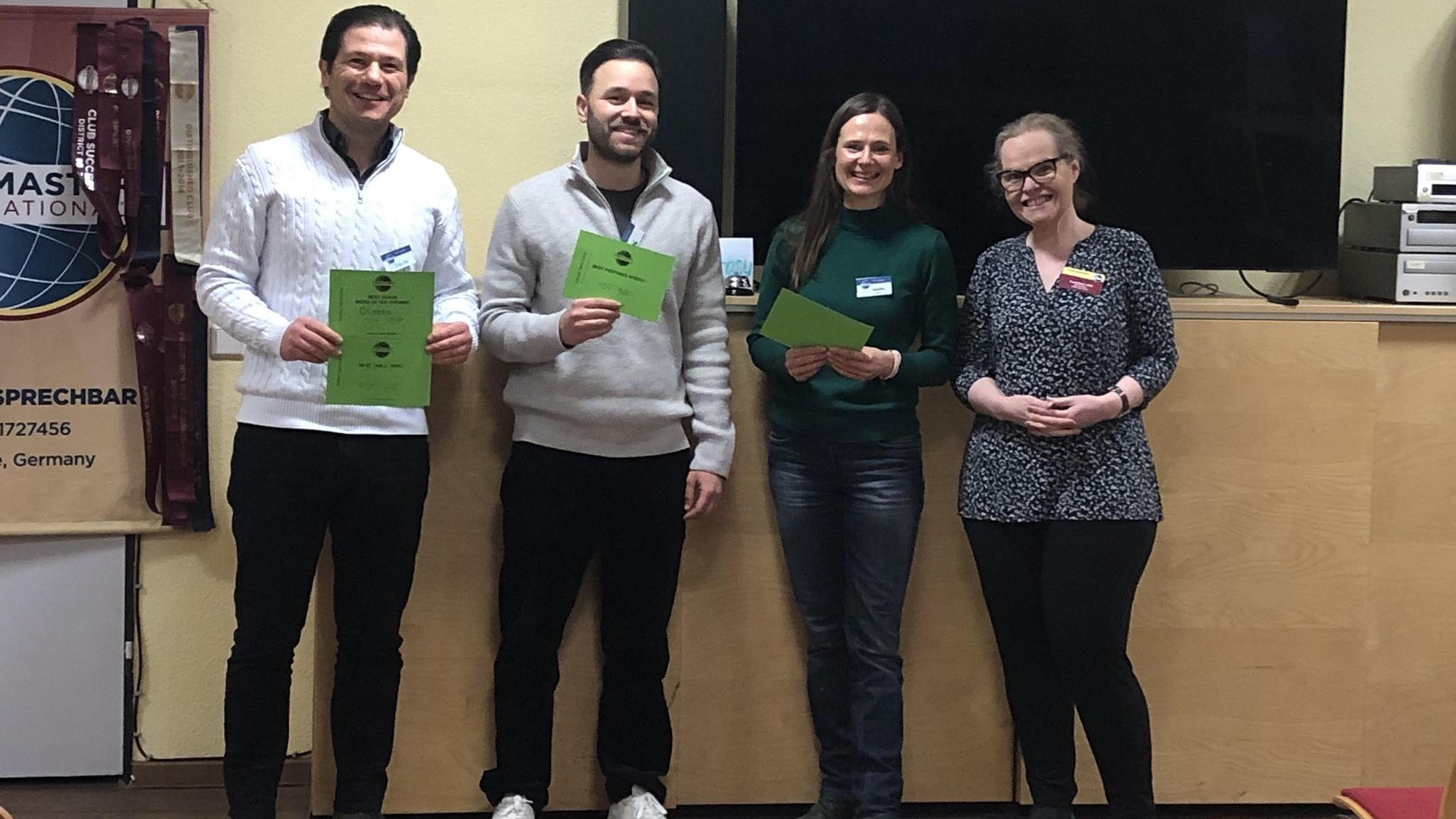 Four members of Kölner Sprechbar smiling with their certificates in front of the Toastmasters banner.