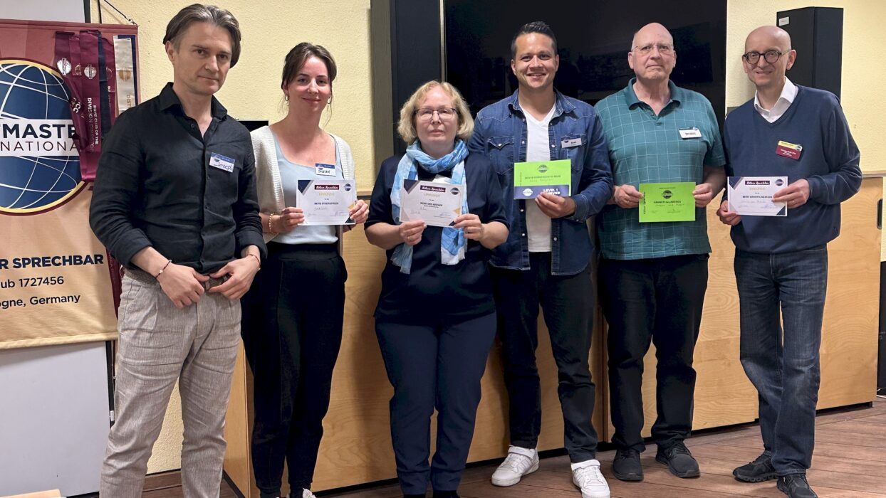 Sechs Mitglieder der Kölner Sprechbar stehen nebeneinander in einem Clubraum, mehrere halten Urkunden in der Hand. Im Hintergrund ein Banner von Toastmasters International und ein Holzschrank mit Schiebetüren.