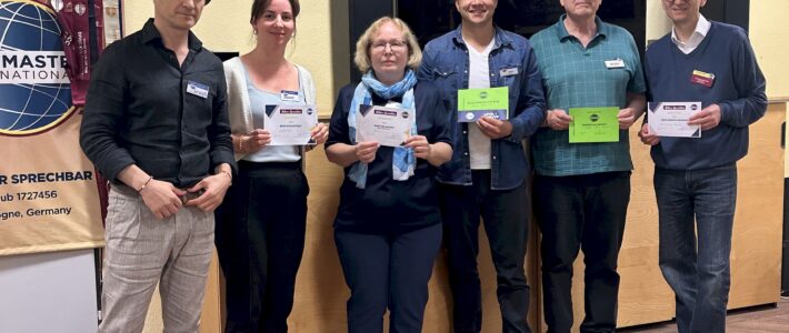 10.06.2026 – Storytelling: Alles vergeht, aber die Geschichten bleiben Sechs Mitglieder der Kölner Sprechbar stehen nebeneinander in einem Clubraum, mehrere halten Urkunden in der Hand. Im Hintergrund ein Banner von Toastmasters International und ein Holzschrank mit Schiebetüren.