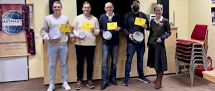 Fünf Toastmasters-Mitglieder stehen lächelnd nebeneinander und halten weiße Teller in den Händen, im Hintergrund ein Fahnenbanner der Kölner Sprechbar.