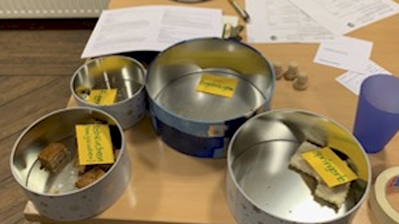 Vier runde Blechdosen mit selbstgebackenen Weihnachtsplätzchen und handbeschrifteten gelben Namensschildchen stehen auf einem Holztisch.
