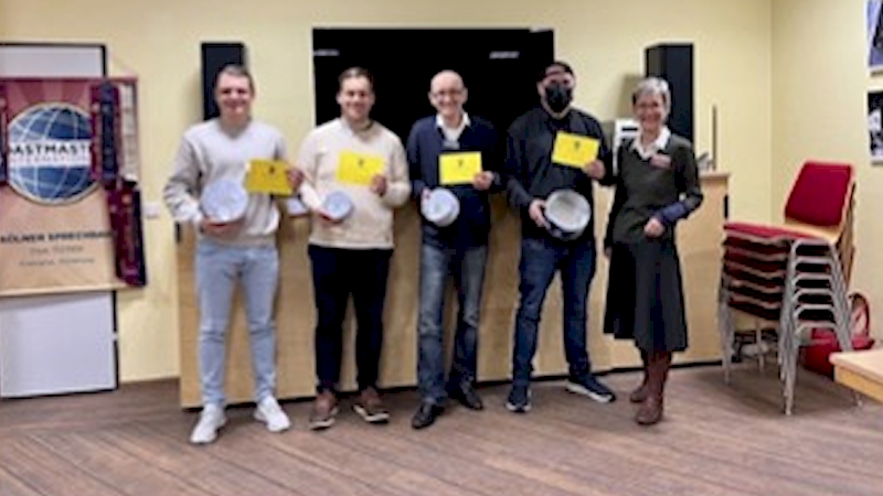 Fünf Toastmasters-Mitglieder stehen lächelnd nebeneinander und halten weiße Teller in den Händen, im Hintergrund ein Fahnenbanner der Kölner Sprechbar.