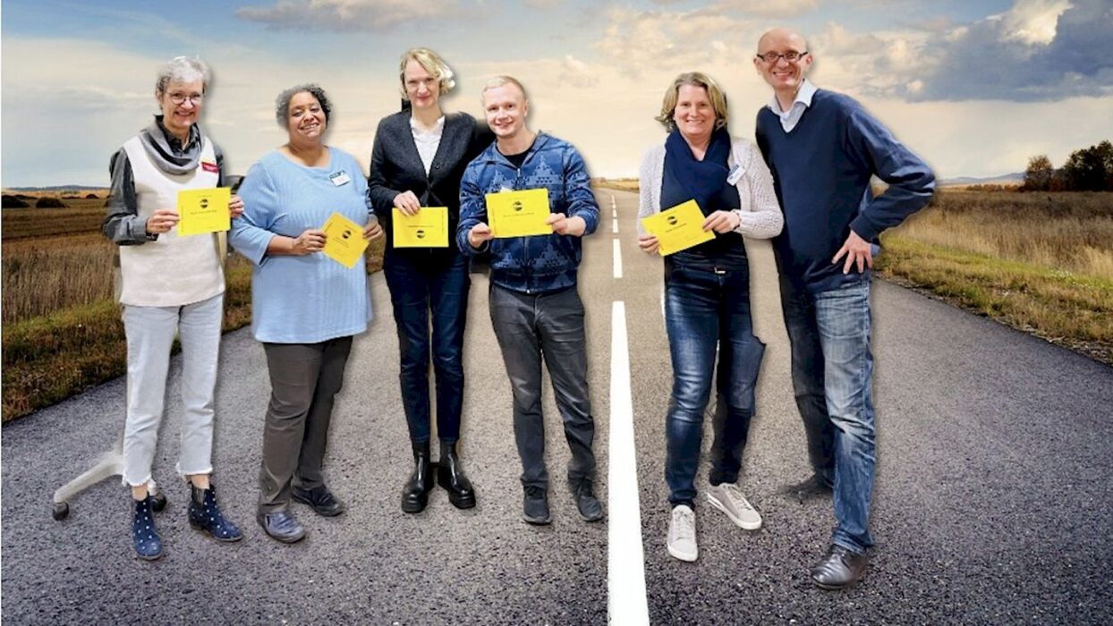 Sechs lächelnde Toastmasters-Mitglieder mit gelben Auszeichnungskarten stehen auf einer Landstraße, im Hintergrund Felder unter weitem Himmel.
