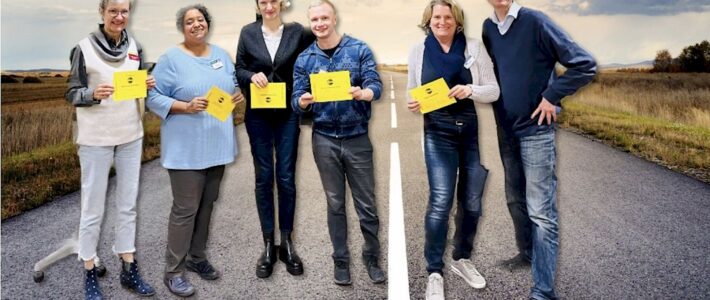 Sechs lächelnde Toastmasters-Mitglieder mit gelben Auszeichnungskarten stehen auf einer Landstraße, im Hintergrund Felder unter weitem Himmel.