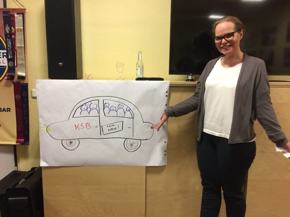 Eine Rednerin steht lächelnd neben ihrem selbstgezeichneten Flipchart-Bild eines Autos mit der Aufschrift "KSB" und "talk.NOW!".