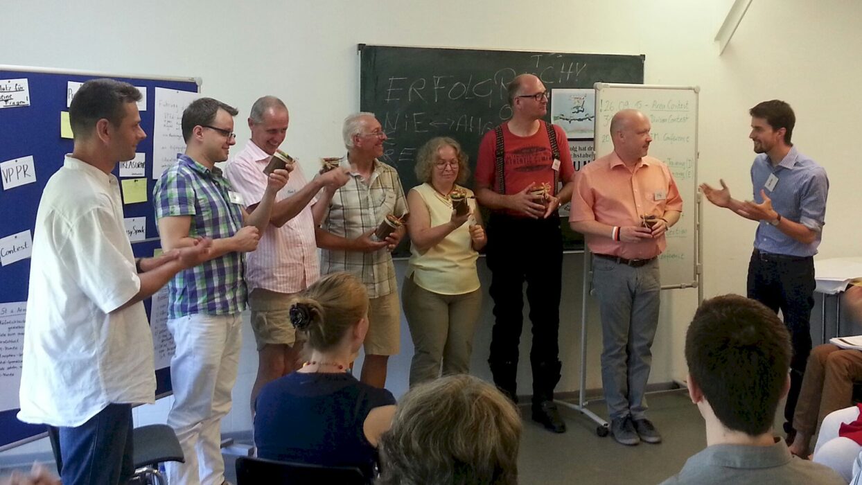 Acht Teilnehmende eines Club Officer Trainings stehen im Stuhlkreis und halten kleine verschnürte Gläser in den Händen, im Hintergrund eine Tafel mit der Aufschrift „Erfolgreich“ und Flipcharts.