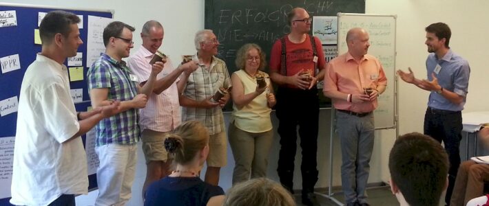 Acht Teilnehmende eines Club Officer Trainings stehen im Stuhlkreis und halten kleine verschnürte Gläser in den Händen, im Hintergrund eine Tafel mit der Aufschrift „Erfolgreich“ und Flipcharts.