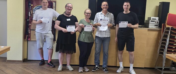 Fünf ausgezeichnete Teilnehmende der Kölner Sprechbar stehen mit Urkunden vor einem Bildschirm im Clubraum.