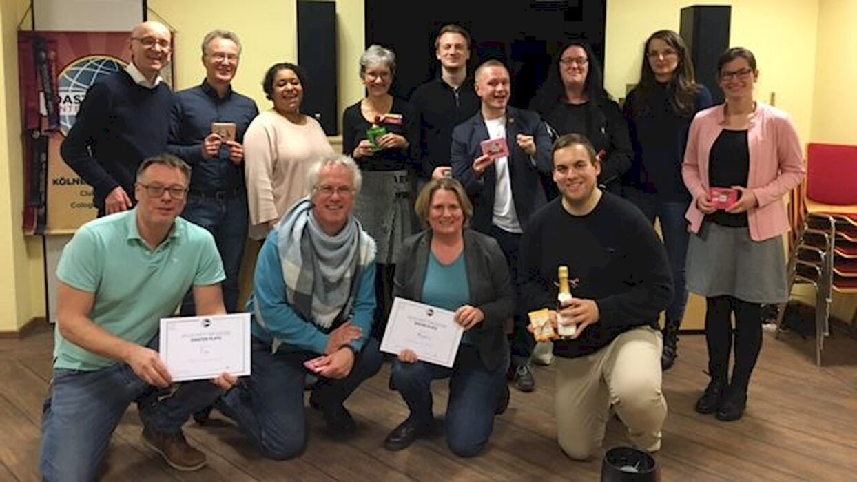 Eine große Gruppe von Toastmasters-Mitgliedern steht und kniet gemeinsam für ein Gruppenfoto, mehrere halten Urkunden und eine Sektflasche in den Händen.