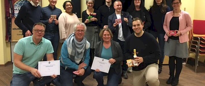 Eine große Gruppe von Toastmasters-Mitgliedern steht und kniet gemeinsam für ein Gruppenfoto, mehrere halten Urkunden und eine Sektflasche in den Händen.
