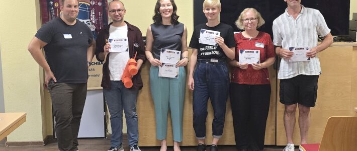 Courage und Geburtstagsfeste bei der Kölner Sprechbar Sechs lächelnde Mitglieder der Kölner Sprechbar stehen nebeneinander vor einem Vereinsbanner und halten ihre Auszeichnungsurkunden in die Kamera; einer von ihnen hält zusätzlich ein selbstgebasteltes rotes Spielzeugtelefon.