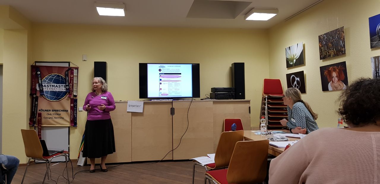 Anne-Kathrin hält einen Pecha-Kucha-Vortrag vor einer Bildschirmpräsentation, im Hintergrund das Toastmasters-Banner der Kölner Sprechbar.