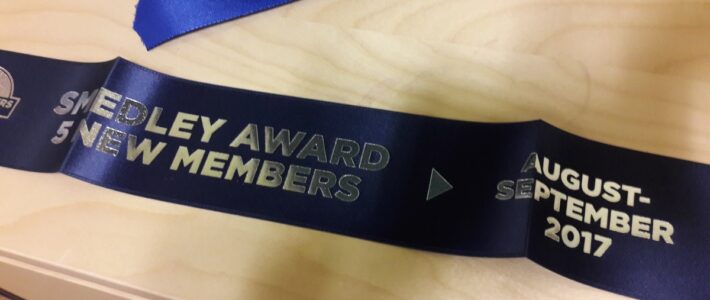 Blaues Wimpelband mit der Aufschrift „Smedley Award 5 New Members August-September 2017“ auf einem Holztisch.