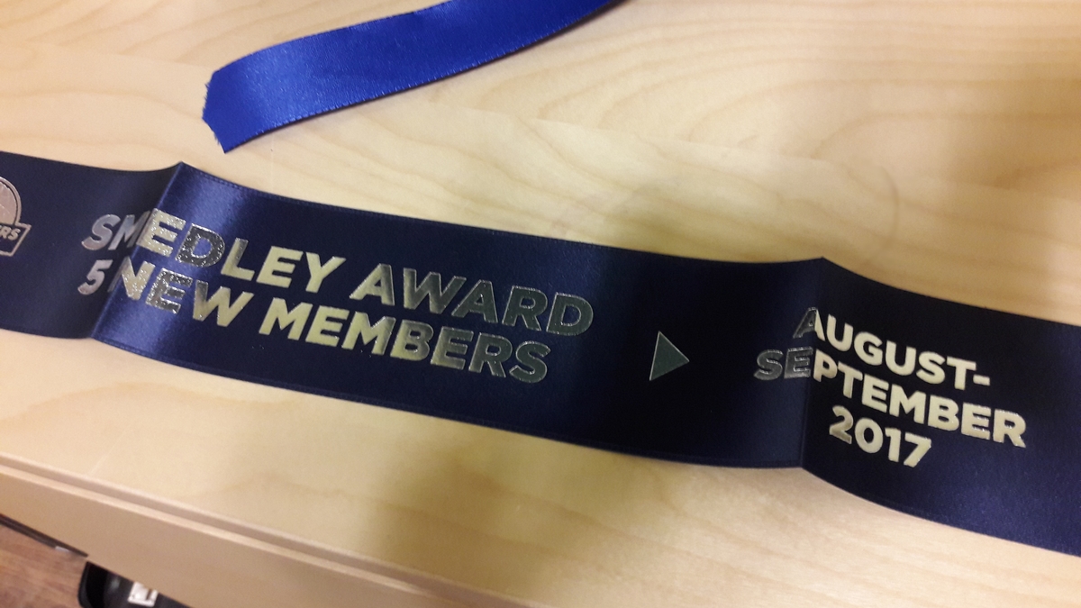 Blaues Wimpelband mit der Aufschrift „Smedley Award 5 New Members August-September 2017“ auf einem Holztisch.