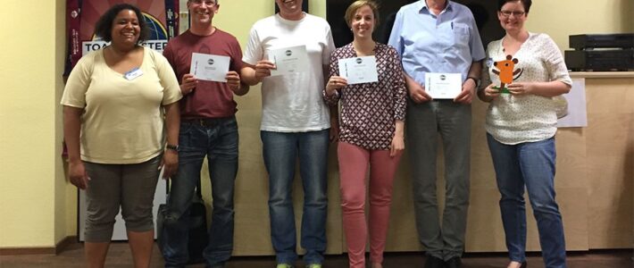 Sechs lächelnde Mitglieder der Kölner Sprechbar stehen nebeneinander vor dem Toastmasters-Banner und halten ihre Urkunden in den Händen.